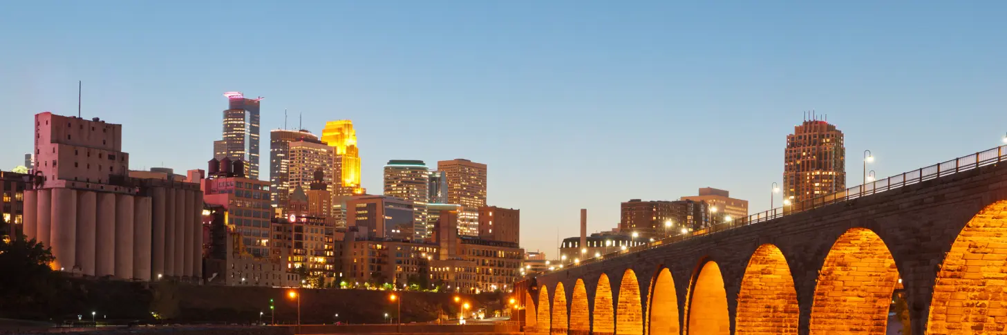 A city skyline at dusk with illuminated skyscrapers and an arched stone bridge.