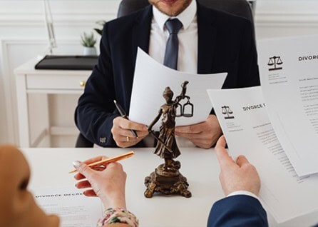 Three individuals in formal attire review divorce documents at a table with a Lady Justice statue in the center.