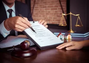 A person signs a contract at a desk with a gavel, scales of justice, and small house models.