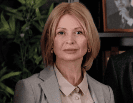 A woman with blonde hair in a gray blazer stands indoors with green plants and a framed photo in the background.