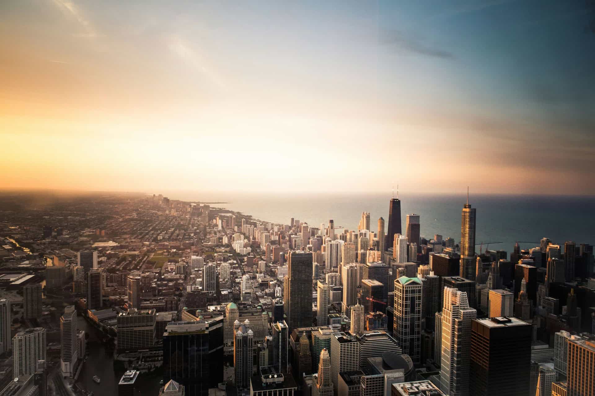 Aerial view of a city skyline at sunset, with tall buildings and a coastline in the background.