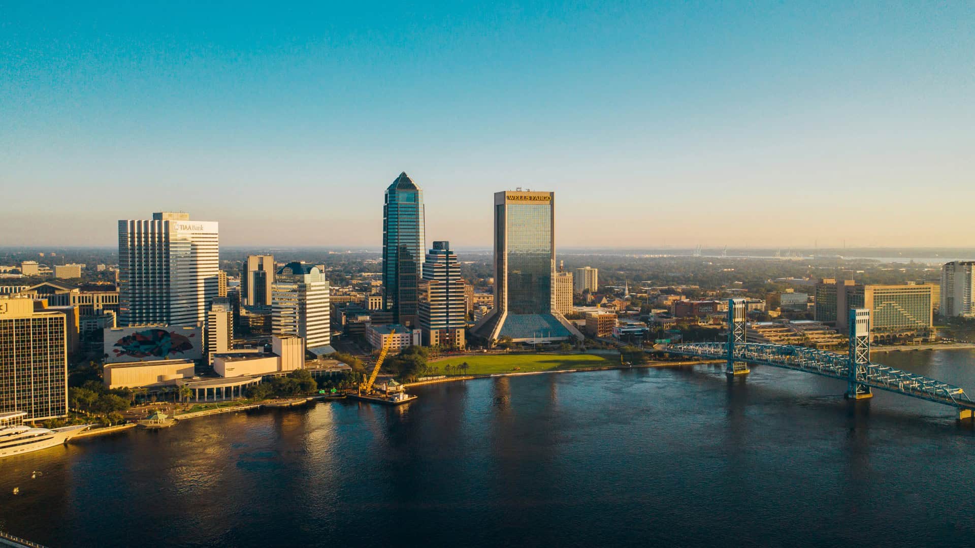 Aerial view of a city skyline with tall buildings, a bridge crossing over a river, and clear blue skies.
