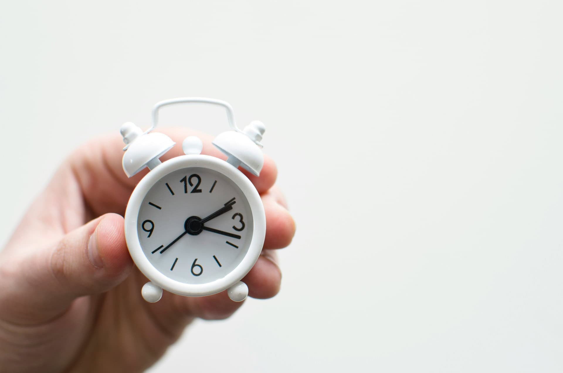 Hand holding a small white analog alarm clock showing 3:00 against a plain background.