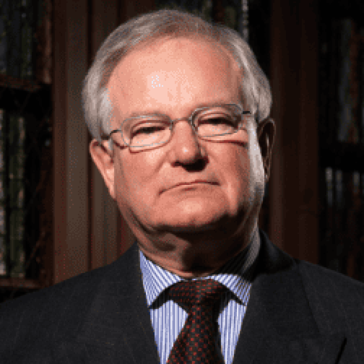 An older man wearing glasses, a suit, and a tie stands in front of bookshelves.